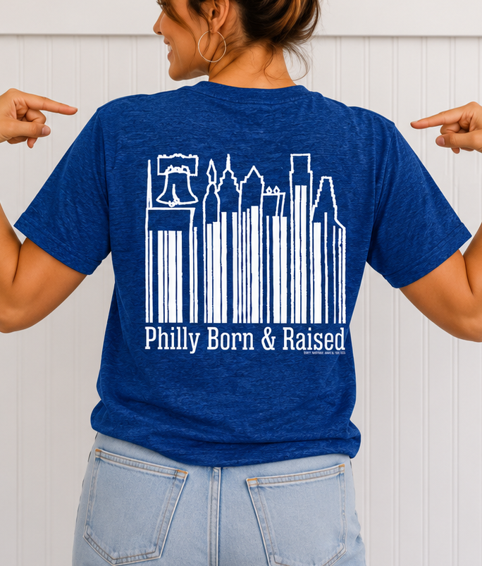 Female model wearing the GOLDENBERG’S® PEANUT CHEWS® Philly Born & Raised Skyline Barcode Tee showcasing the large Philadelphia skyline barcode graphic on the back of a royal blue unisex T-shirt.