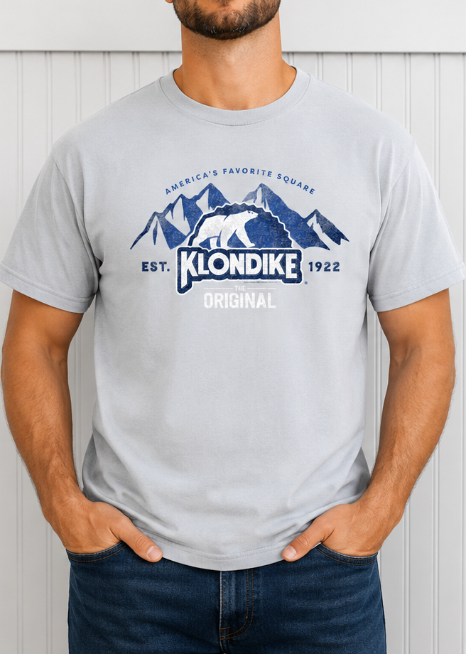 Male model wearing a Klondike® The Original Since 1922 Vintage Unisex Graphic Tee in light gray featuring the iconic polar bear logo and mountain-inspired retro design.