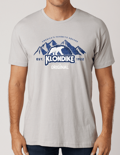 Male model wearing a Klondike® The Original Since 1922 Vintage Unisex Graphic Tee in light gray featuring the iconic polar bear logo and mountain-inspired retro design.